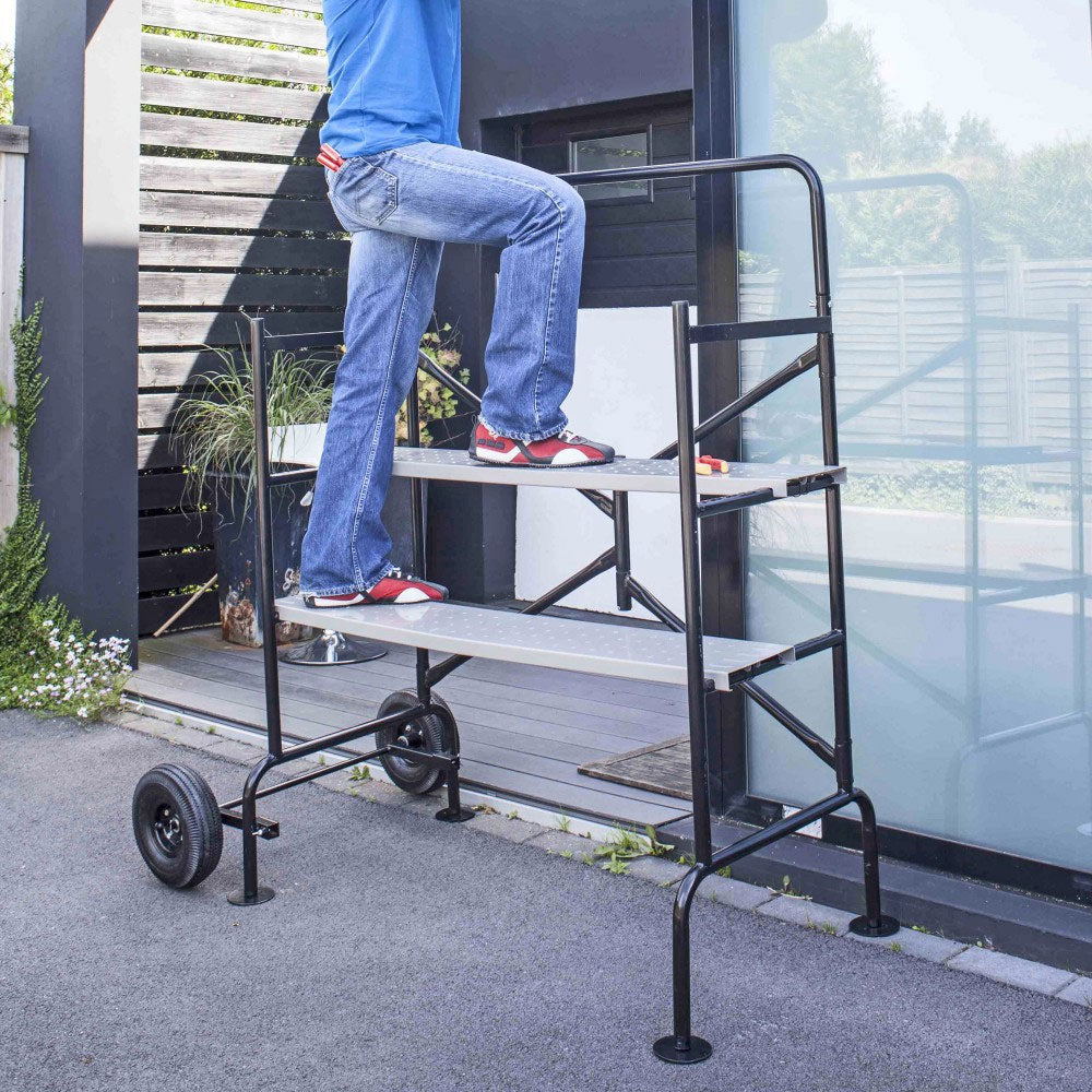 Mobiles Arbeitsgerüst mit zwei Stufen, schwarzer Metallrahmen und zwei großen Rädern, Sitzfläche aus Holz.